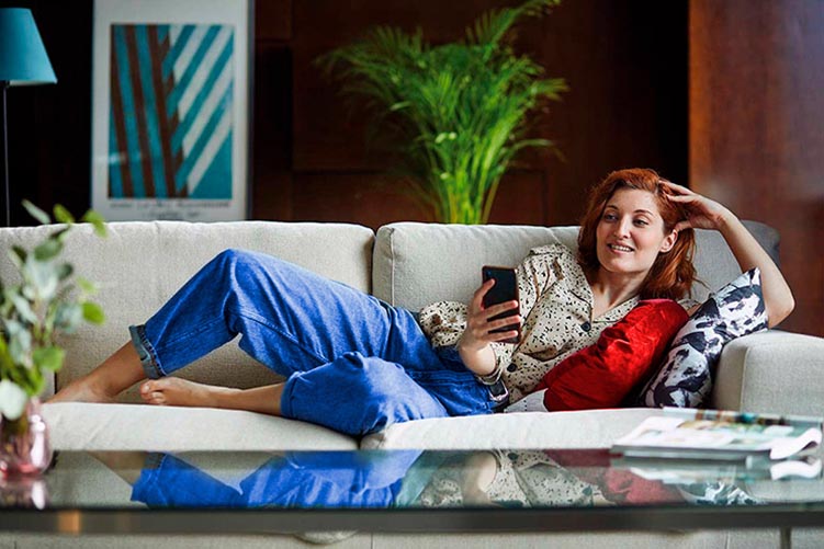 Mujer tumbada en el sofá de su segunda residencia mientras revisa su teléfono móvil
