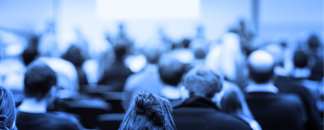 Imagen en tonos azules de una sala de congresos con personas atendiendo a los ponentes de una mesa redonda.