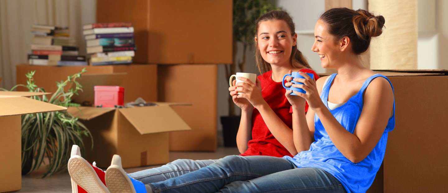 Imagen dos chicas sentadas en el suelo tomando café rodeadas de cajas de mudanza