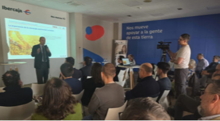 Sala de conferencia con pantalla proyectando una presentación. El ponente interviene y el público les está escuchando.