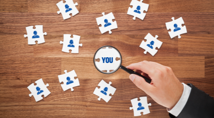 Piezas de puzzle con la representación de personas y una mano con una lupa destacando la pieza central en la que pone You